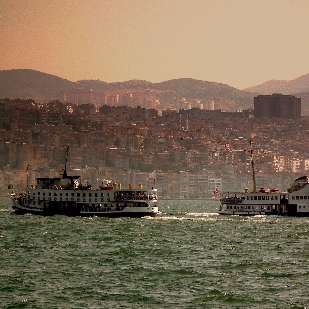 Under a hazy sky, two ferries glide across the waters, with a bustling cityscape and majestic mountains in the backdrop.