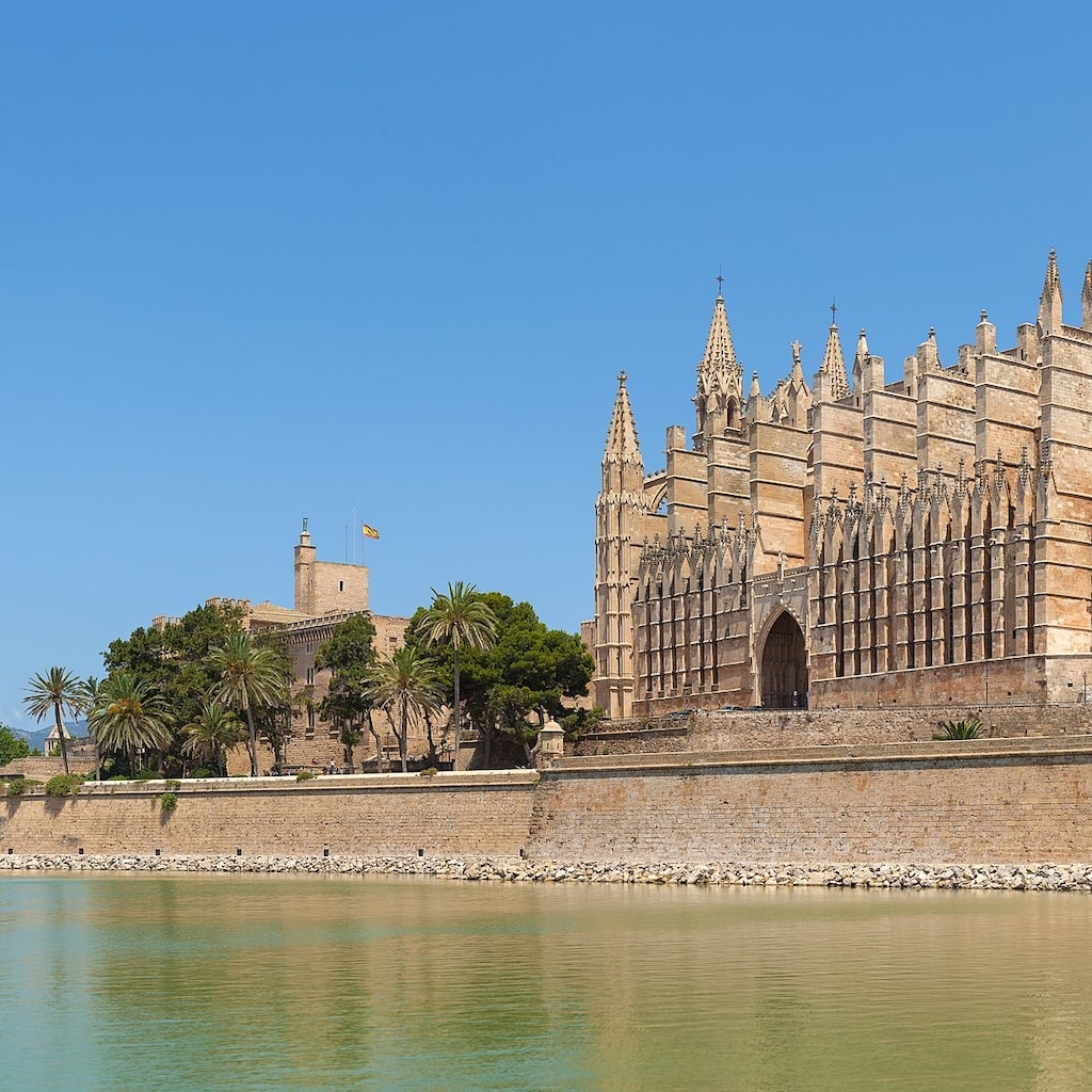 A grand Gothic cathedral, adorned with ornate spires, stands majestically by a tree-lined waterfront under a crystal-clear sky.